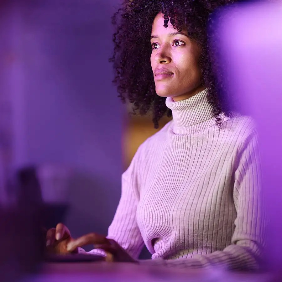 A woman in a ribbed turtleneck sweater works at a computer, bathed in striking purple and blue lighting.