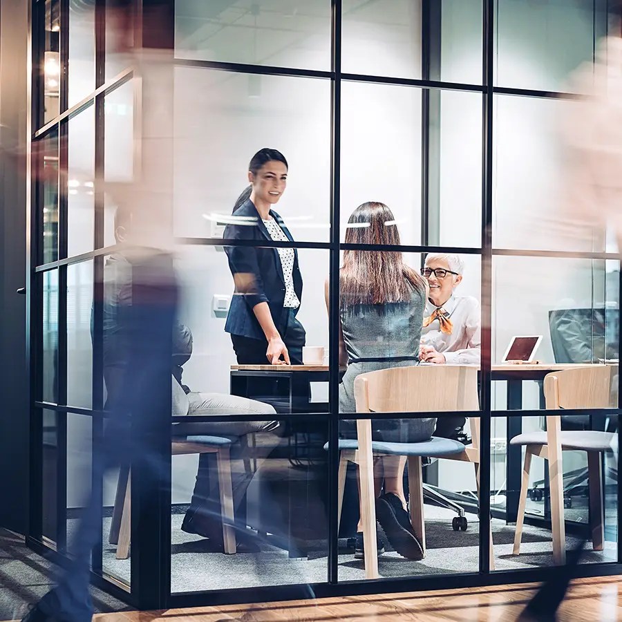 Three women hold a meeting in a glass-walled office at an Appeals Management Services firm, creating a dynamic workspace scene.