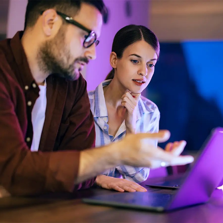 Two people at a table study a laptop, discussing Analytics-as-a-Service, under vibrant purple lighting.