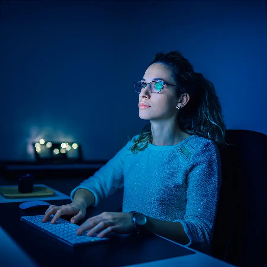 Woman wearing glasses works at a computer, reflecting How We Deliver in a blue-lit, dimly lit room with blurred lights behind.