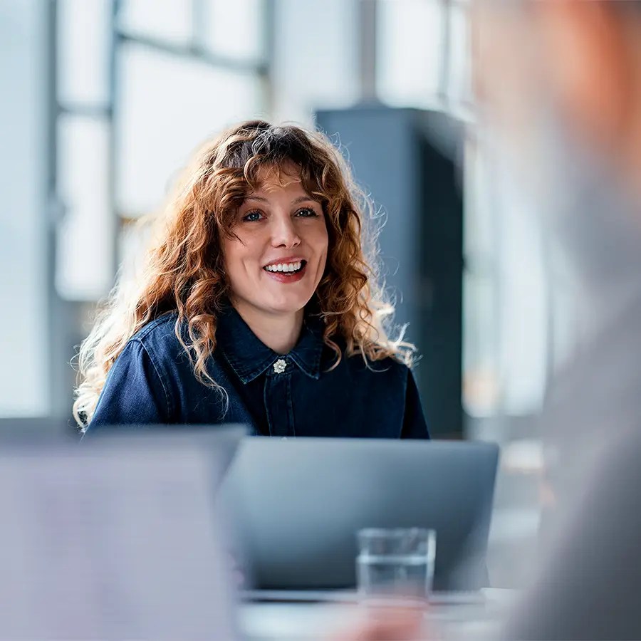 Smiling woman with curly hair sits at a desk using Cloud Analytics on her laptop in a bright, modern office setting.