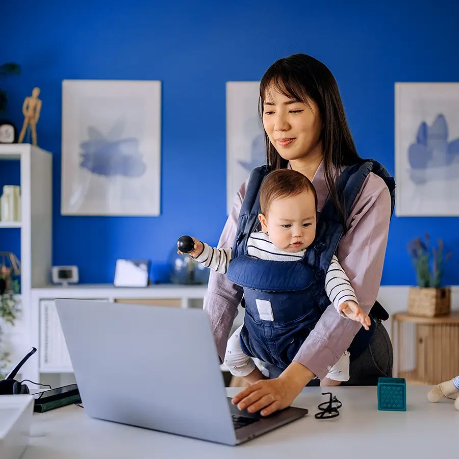 Woman working on a laptop with a baby in a carrier in a modern, blue home office, representing patient access,