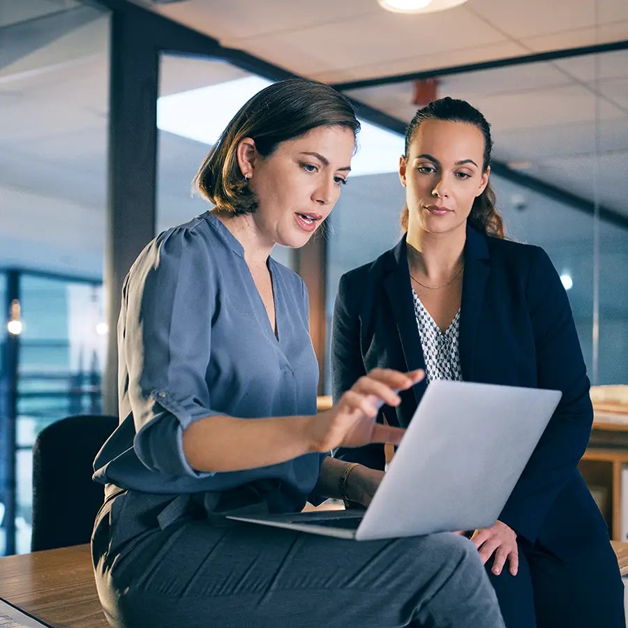 Two women in business attire discuss Product & Plan Configuration services on a laptop in a modern glass-walled office.