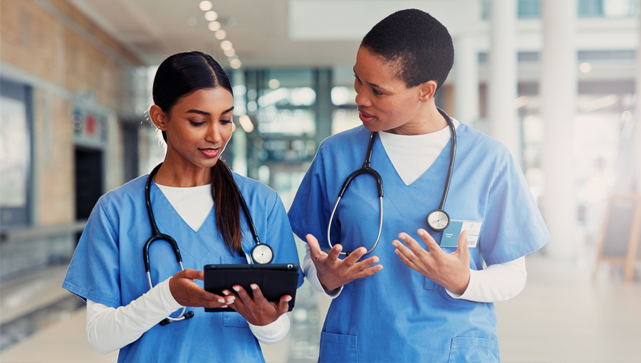 Two healthcare workers in scrubs discuss from a clinical perspective while looking at a tablet in a bright hospital corridor.