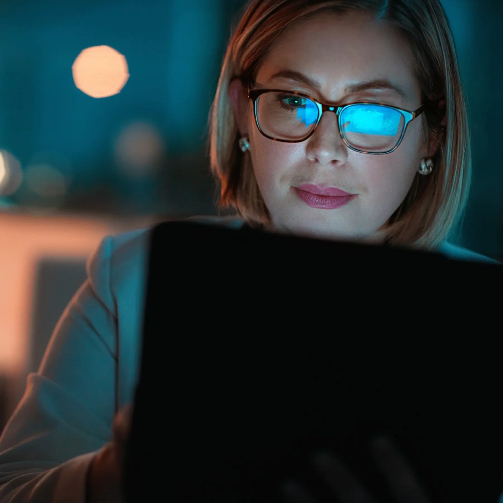 Woman with glasses using a SmarTec Nurse Assist device, tablet screen light reflecting on her glasses in a dimly lit room.