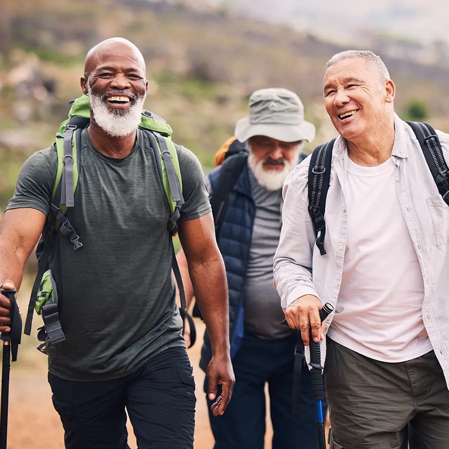 Three older men, smiling, hike outdoors on a trail, showcasing sagility and synchrony amid lush greenery.
