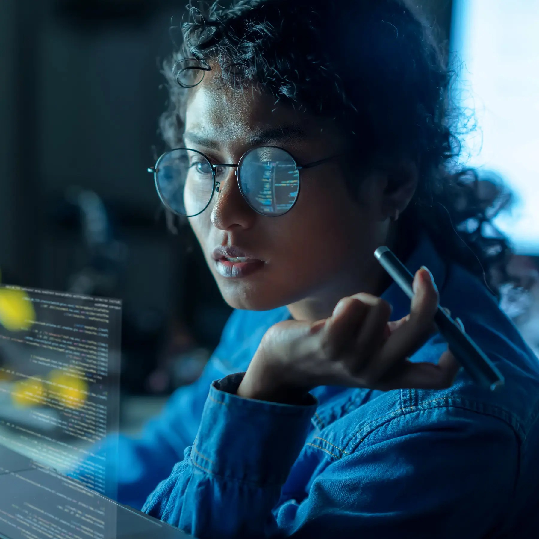Person with curly hair and glasses reviewing Nurse Assist code on a screen, pen in hand, code reflected in glasses.