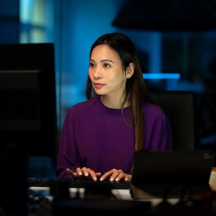 A woman in a purple top works at a computer on Integrated Partner Operations in a dimly lit, blue-lit office.
