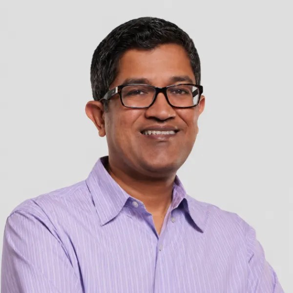 Smiling man with short dark hair and glasses, wearing a light purple button-up shirt, stands against a gray background.