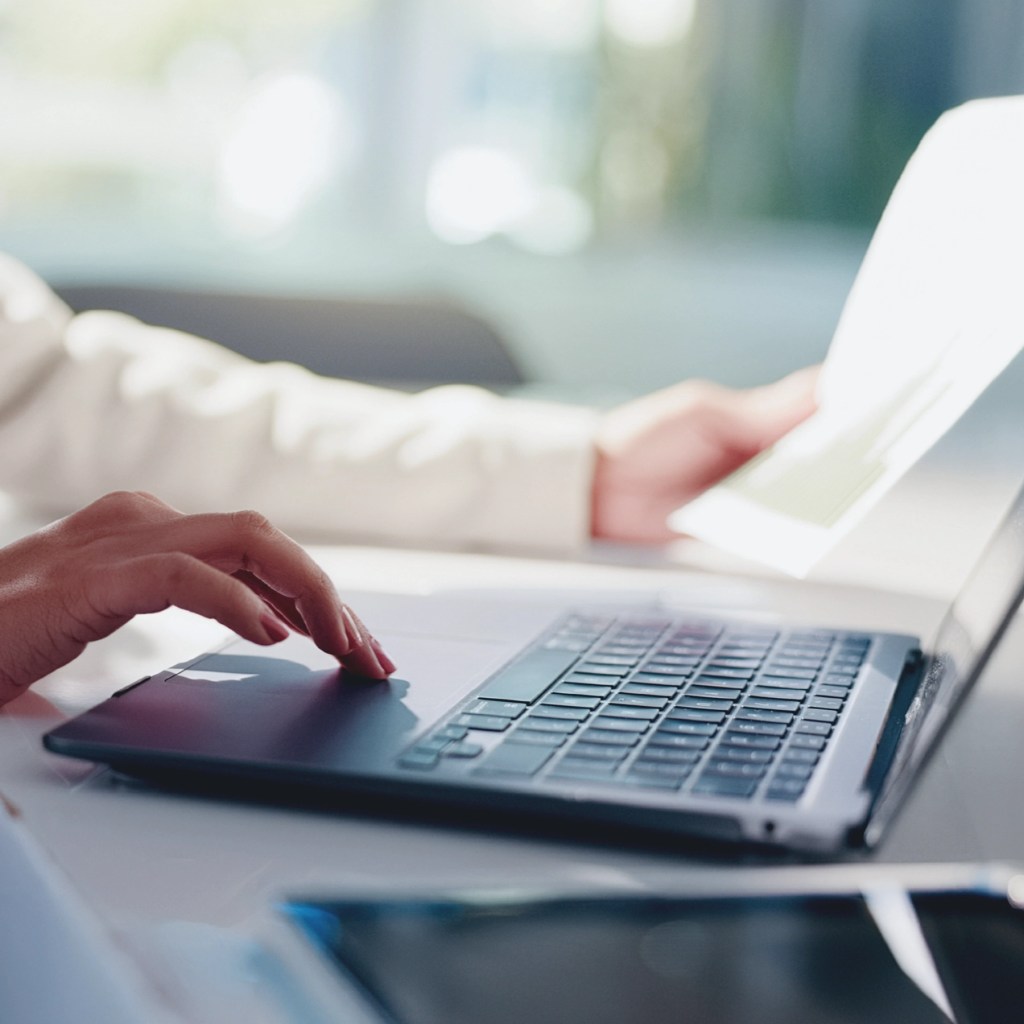 A person uses a laptop touchpad with one hand while holding HEDIS Stars Performance sheets at a bright desk.
